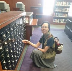 person with black hair tied back and wire glasses, looking at the camera, squatting next to a card catalog drawer set, reaching out to open a drawer in the Enoch Pratt Baltimore library during the daytime.