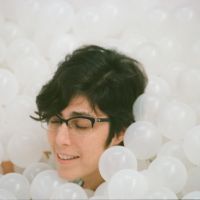 A white woman with short brown hair and glasses' face emerging from a sea of white plastic balls with her eyes closed.