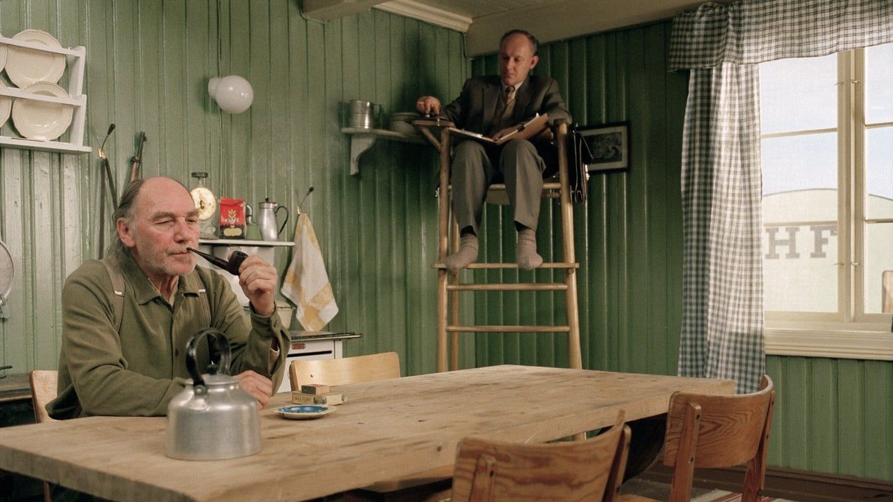 still from the film Kitchen Stories. A white man with a grey beard sits at a kitchen table smoking a pipe. In the corner of the room, another man sits in a tall lifeguard-style chair observing the other man. The man in the chair is writing in a notebook.